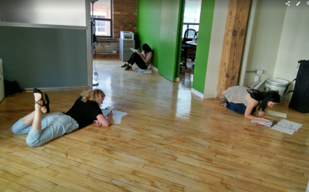 Office space with hardwood floors and green walls. Three young women are either lying, sitting or kneeling on the floor, each sketching on multiple sheets of paper.