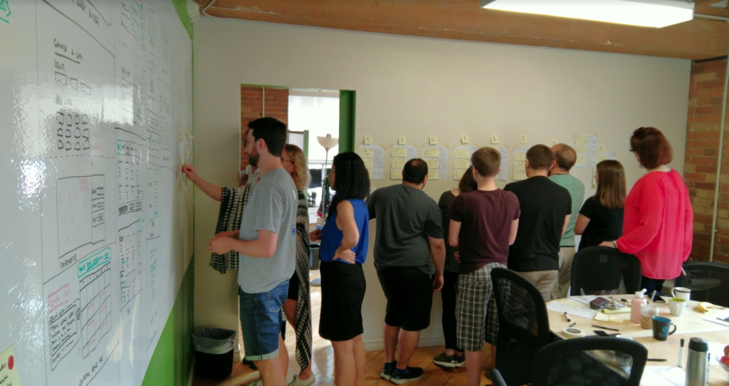 A group of casually dressed people focusing on drawings and hung sheets of paper on two adjoining walls. No faces visible.